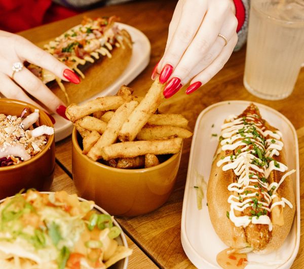 mains d'une femme en train de prendre des frites, avec des hot-dogs, des nachos, et une citronnade autour.